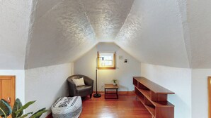 A cozy attic room with a sloped ceiling, a single window, a leather armchair, a small table with a lamp, a wooden bench, and a patterned ottoman. - Snap, Set, Stay- Game Day Comfort Near It All (Canton)