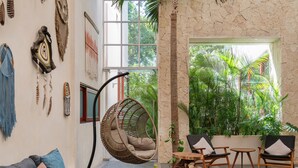 A cozy living area with a sofa, a hanging chair, and a small table. There are plants and a large window in the background. - WAYAK Luxury! 5BR Private Pool & Gym, 24/7Security (Tulum)