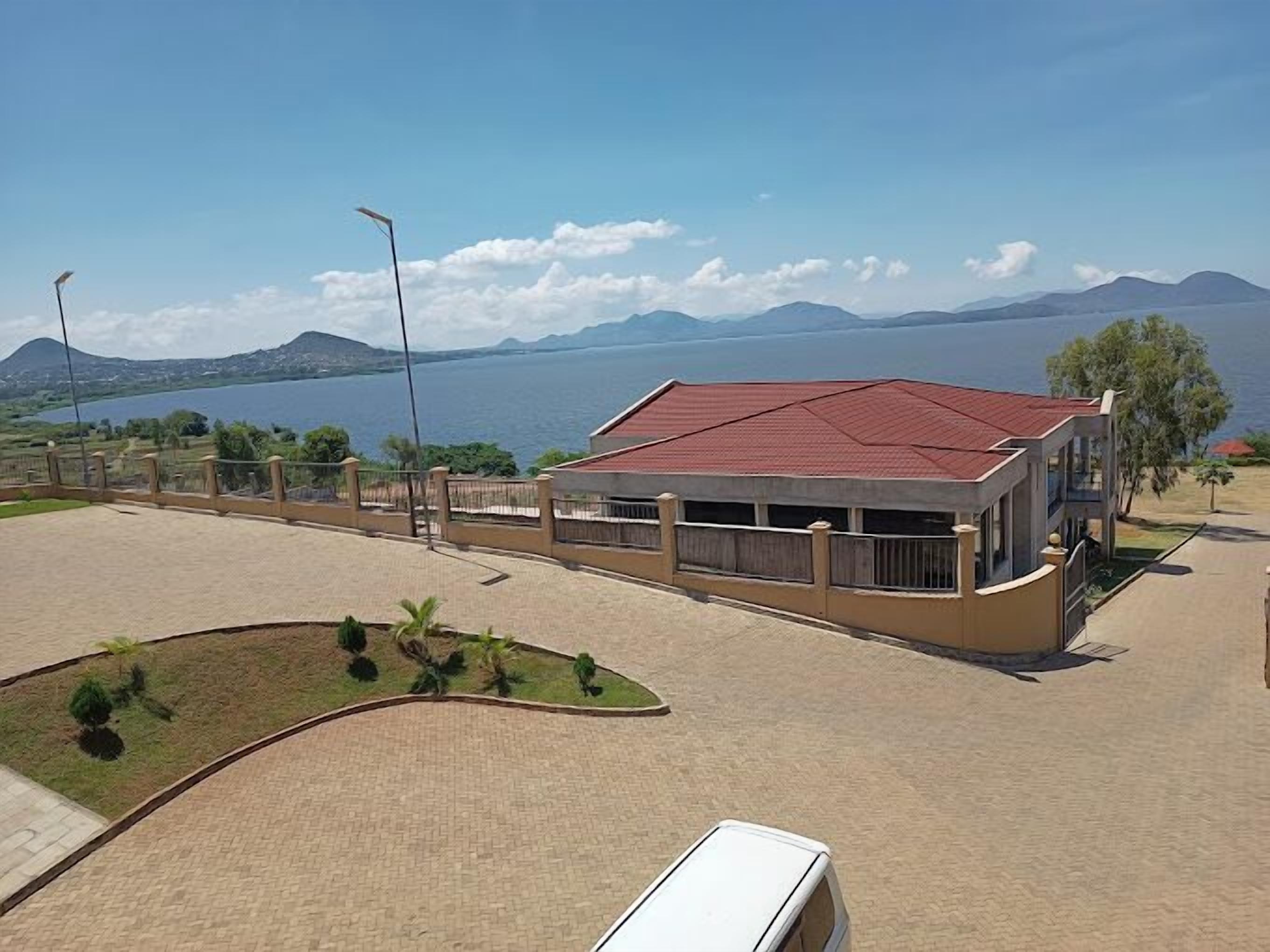 Um edifício com telhado vermelho, área pavimentada na frente e vista para um corpo de água e montanhas ao fundo.