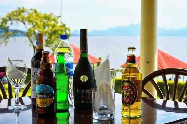 A table with various bottles and glasses, likely containing beverages, set against a scenic backdrop with a body of water and mountains. - Golden Rays Resort (Homa Bay)