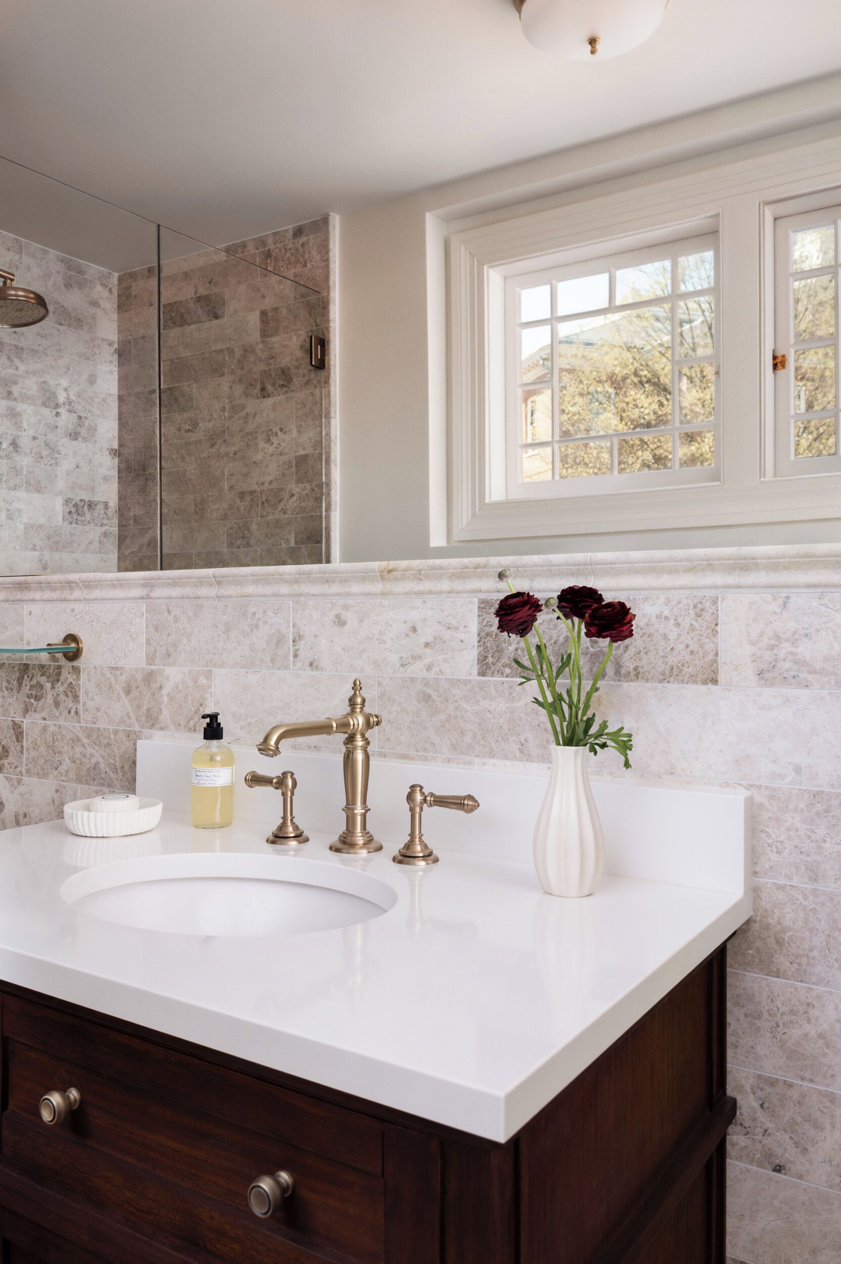 A bathroom with a white sink, brass faucets, a vase with flowers, and a window with a view of trees.