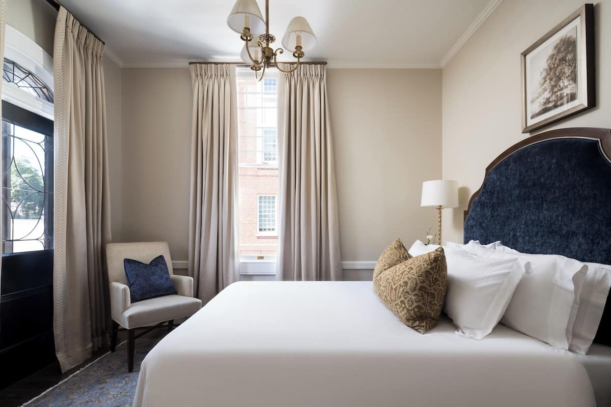Une chambre d’hôtel avec un grand lit, une chaise, un lustre et une fenêtre avec des rideaux.