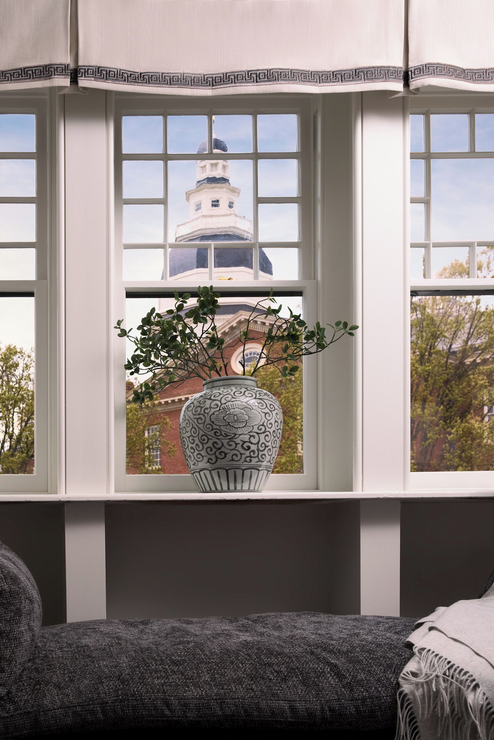 A room with a large window featuring a decorative white vase with a plant, a view of a building with a dome, and a grey upholstered bench.
