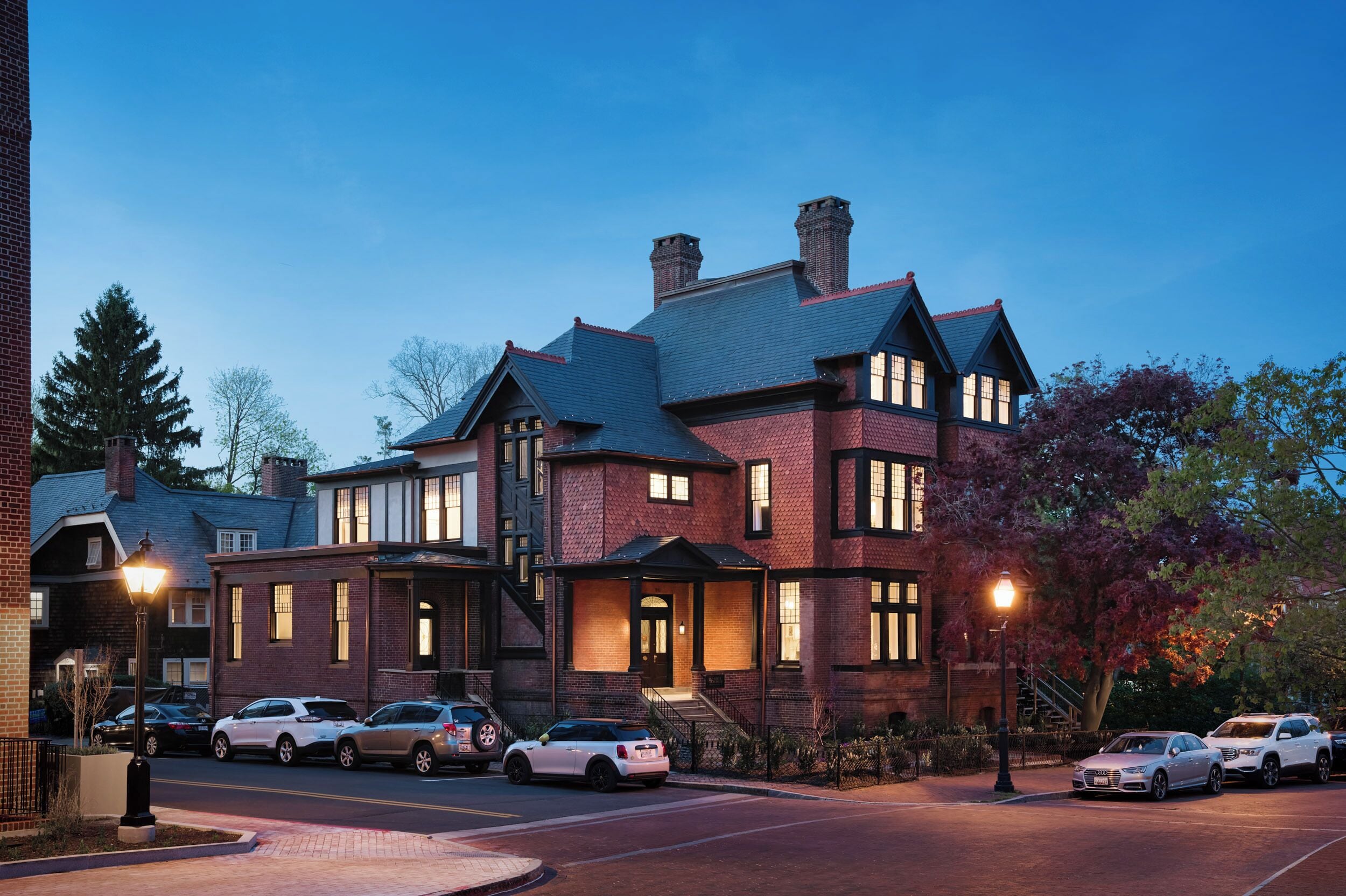 A large brick building with multiple windows and a prominent chimney, surrounded by parked cars and trees.