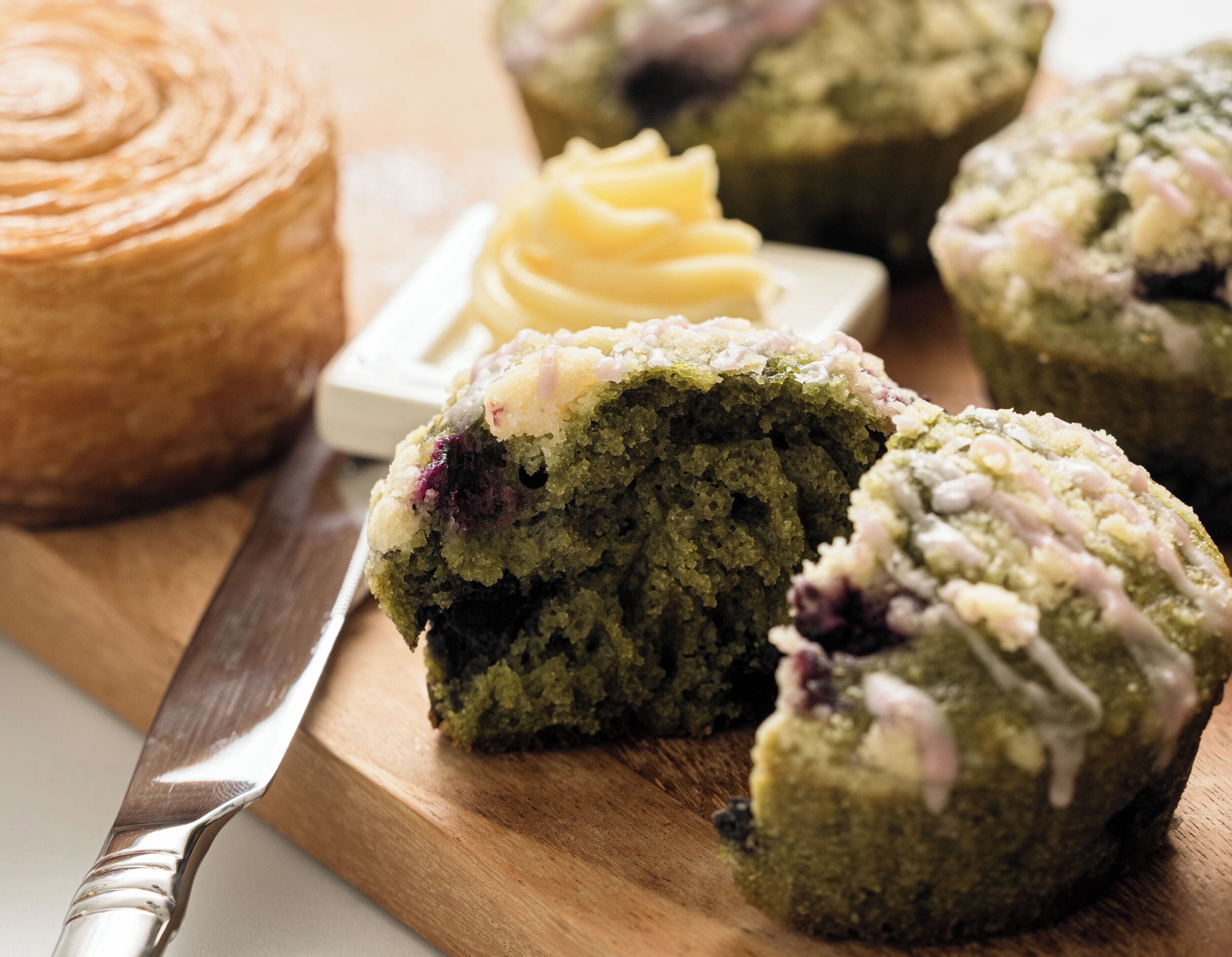 A wooden cutting board with a green muffin, a knife, and a container of butter.