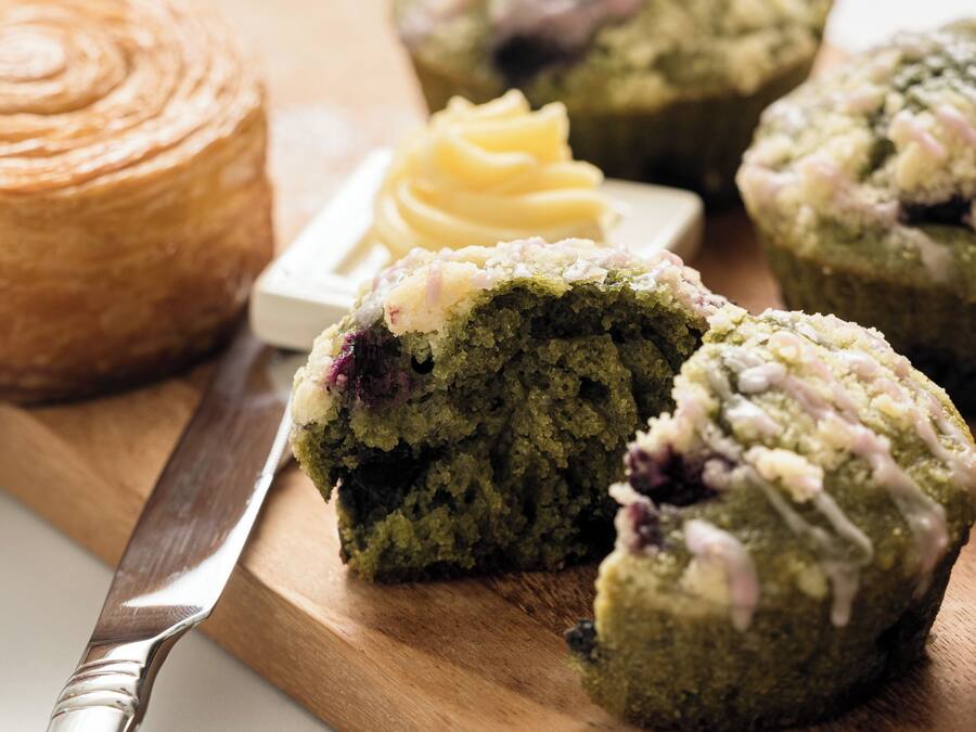 A wooden cutting board with a green muffin, a knife, and a container of butter.