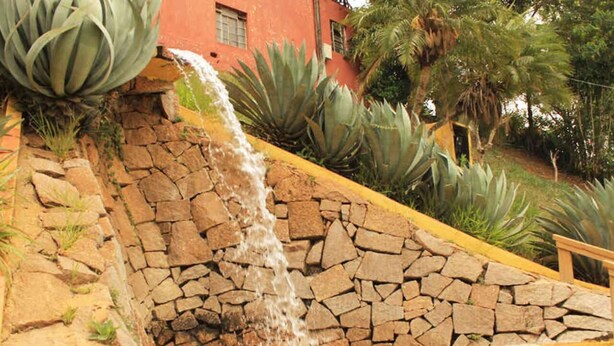Uma fonte de pedra com cascata, rodeada por grandes plantas de agave e um edifício vermelho ao fundo.