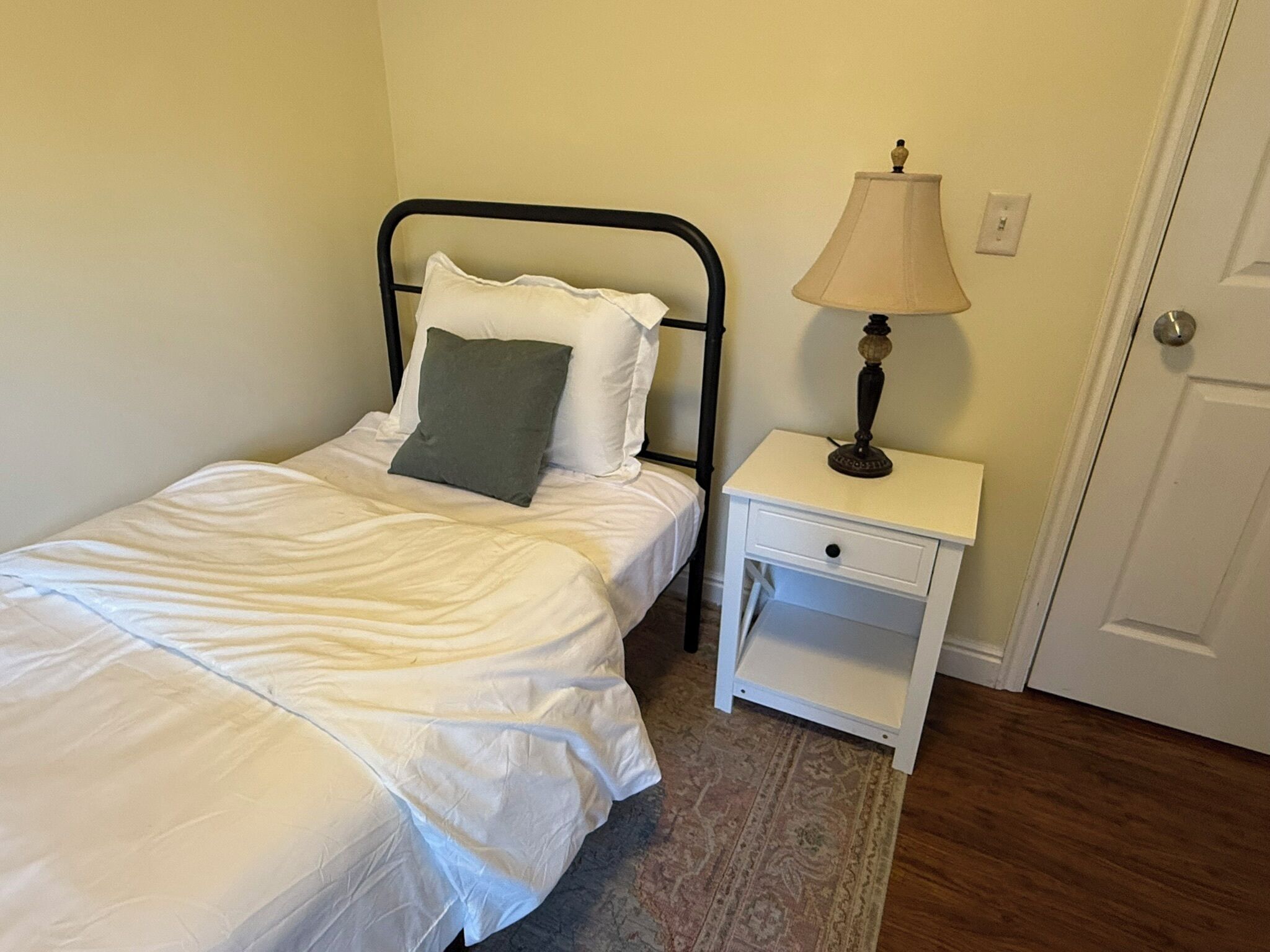 Una cama individual con ropa de cama blanca, una almohada verde, una mesita de noche con lámpara y una puerta al fondo.