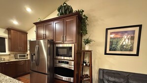 A kitchen with a refrigerator, microwave, oven, and wooden cabinets. There is a plant on a shelf, a framed picture on the wall, and a leather couch in the living area. - Heber Valley Base Camp: convenient location, modern amenities, pleasant spaces (Heber City)