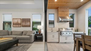 A modern living room with a sofa, a coffee table, and a dining area with a wooden table and chairs. A kitchen with a stove and cabinets is visible through an open door. - LUXE: Custom Beach Home w/Pool&Spa, Fire Pit, BBQ (Naples)