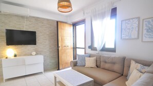 Una sala de estar moderna con un sofá, un televisor montado en la pared, una puerta de madera y un ventanal con cortinas.