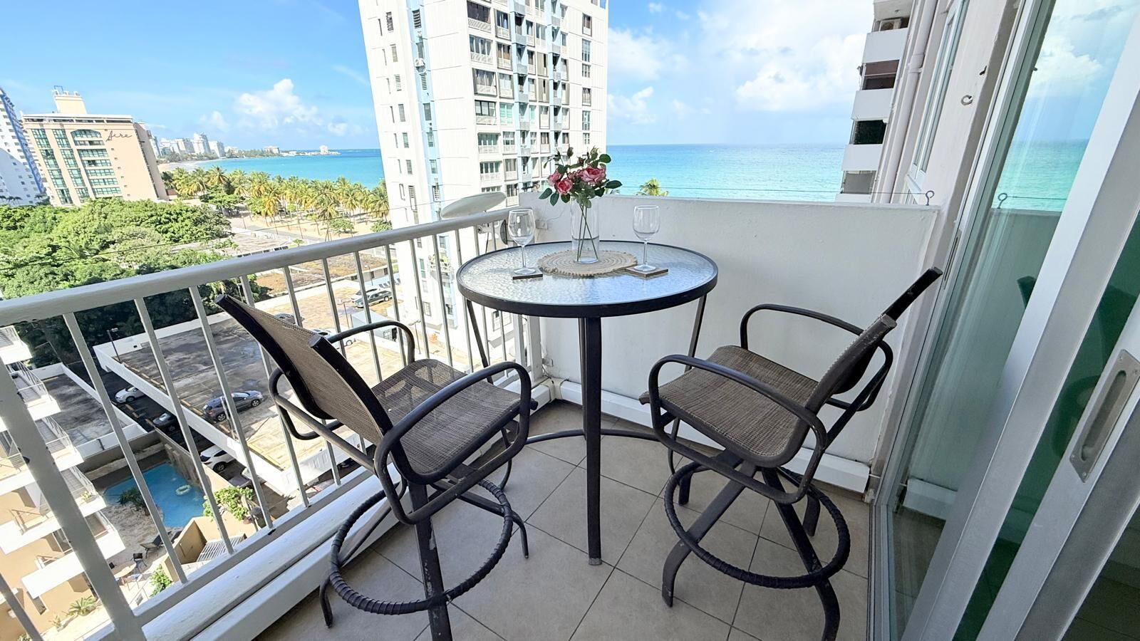 A balcony with two wicker chairs and a small round table, overlooking a pool and a beach.