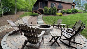 Un espace extérieur circulaire aménagé avec des chaises Adirondack et des tables de pique-nique, entouré d’un foyer en pierre, devant une maison en bois dotée d’un porche.