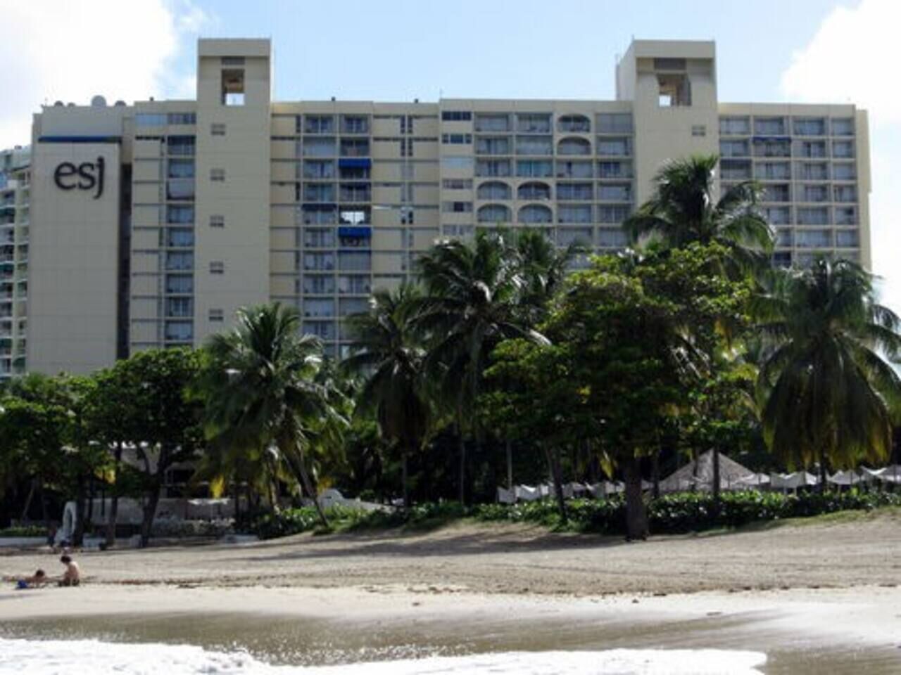 A hotel with multiple floors, palm trees in the foreground, and a beach in the background.