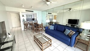 A living room with a blue sofa, glass coffee table, and wicker chairs. There is a kitchen in the background with white cabinets and a dining area with chairs.