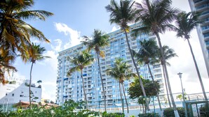 Un hotel alto con varios balcones, rodeado de palmeras y una zona delimitada por una cerca.