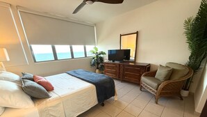 Un dormitorio con cama, televisor, una silla y un ventanal amplio.