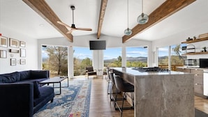 A spacious living area with a dark sofa, a coffee table, and a large rug. There is a kitchen area with a bar and a TV mounted on the wall. - The Sanctuary at Prescott Heights |Majestic Views |Luxe Prescott Sanctuary |Golf (Prescott)