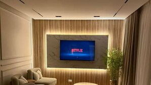 A modern living room with a large flat-screen TV displaying the Netflix logo, a plush sectional sofa, a small round coffee table, and a potted plant.