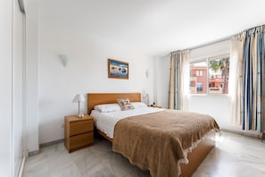 Quarto com cama de madeira, roupa de cama branca, mesa de cabeceira, abajur, um quadro na parede e janela com cortinas.