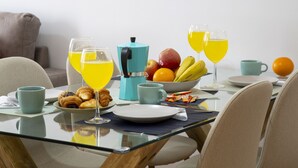 Un desayuno con cafetera, croissants, fruta y vasos de zumo de naranja sobre una mesa de vidrio.