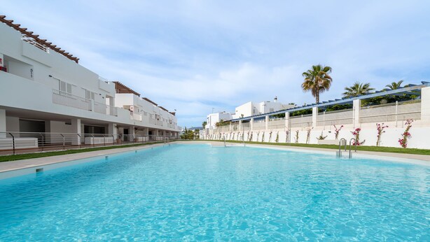 Una piscina con superficie de agua azul cristalina, rodeada de edificios blancos con balcones y una palmera al fondo.