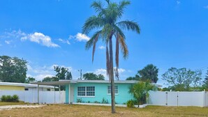Una casa de un piso con fachada turquesa, cerca una valla blanca y un palmera en el jardín.