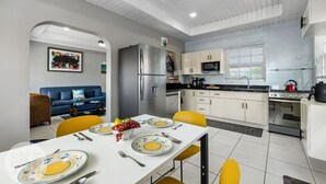A modern kitchen with white cabinets, a stainless steel refrigerator, a microwave, and a stove. There is a dining table with yellow chairs and a white tablecloth. - Cinderella Oasis (Noord)