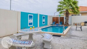 A swimming pool with a blue and white tiled deck, two outdoor tables, and a palm tree. - Cinderella Oasis (Noord)