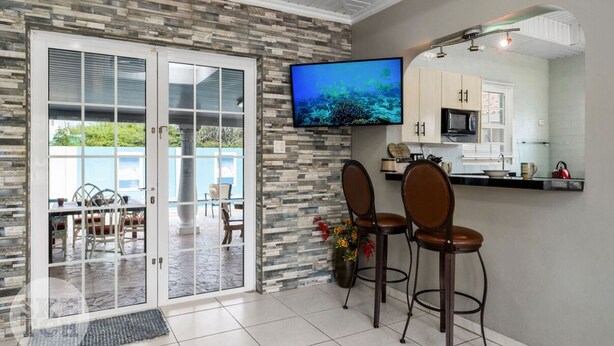 A modern kitchen with a flat-screen TV, a dining area with a glass door leading to a patio, and a stone accent wall. - Cinderella Oasis (Noord)