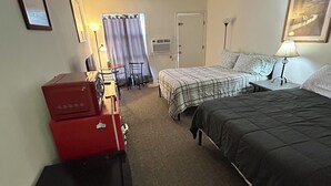 A hotel room with two beds, a desk with a lamp, a chair, a red suitcase, and a door.
