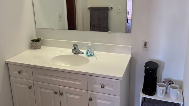A bathroom with a white vanity, a mirror, a sink, a faucet, a soap dispenser, a towel rack, a coffee maker, and a shelf with towels.