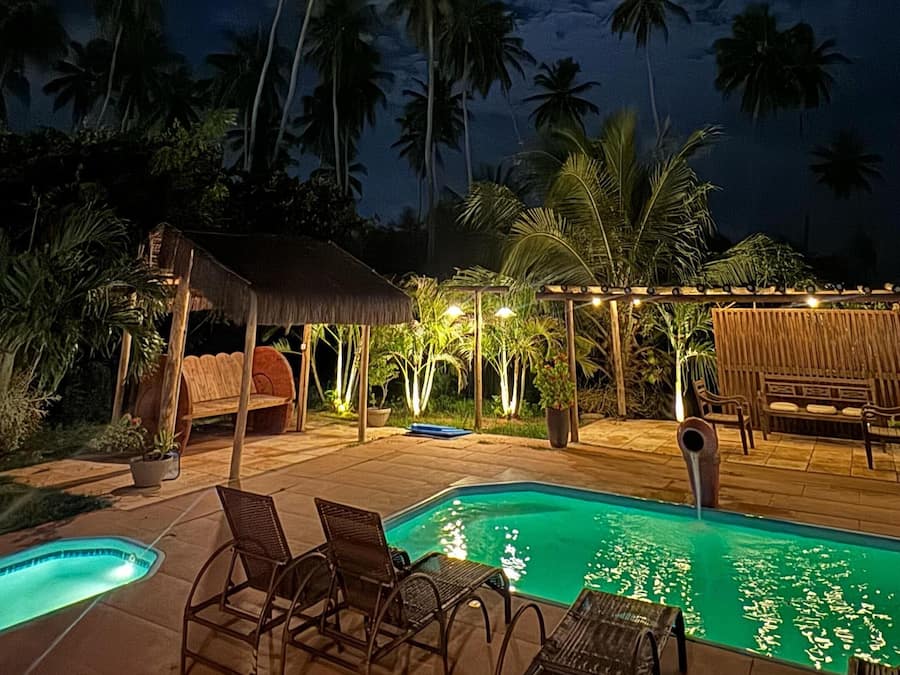 Un área de piscina nocturna con sillones de descanso y una estructura de techo de paja.