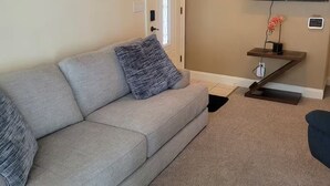 A living room with a grey sofa, a small wooden side table with a vase, a flat-screen TV mounted on the wall, and a door with a decorative glass panel.