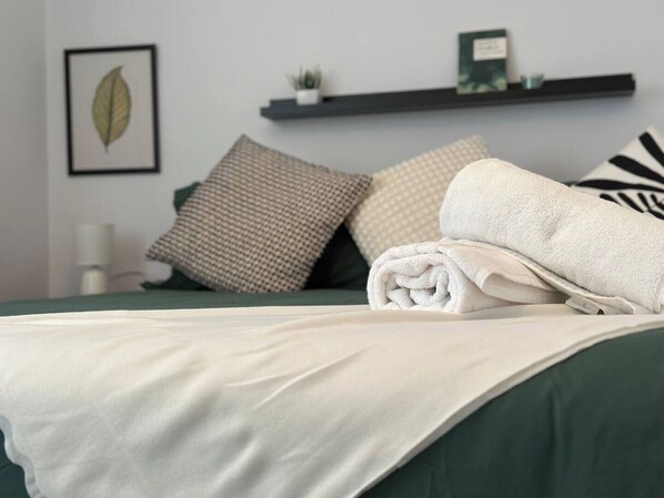 A neatly made bed with a green bedspread and white linens, a small plant on a shelf, and a framed leaf artwork on the wall.