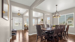 A spacious dining area with a wooden table and chairs, a chandelier, and a view of the outdoors through large windows.