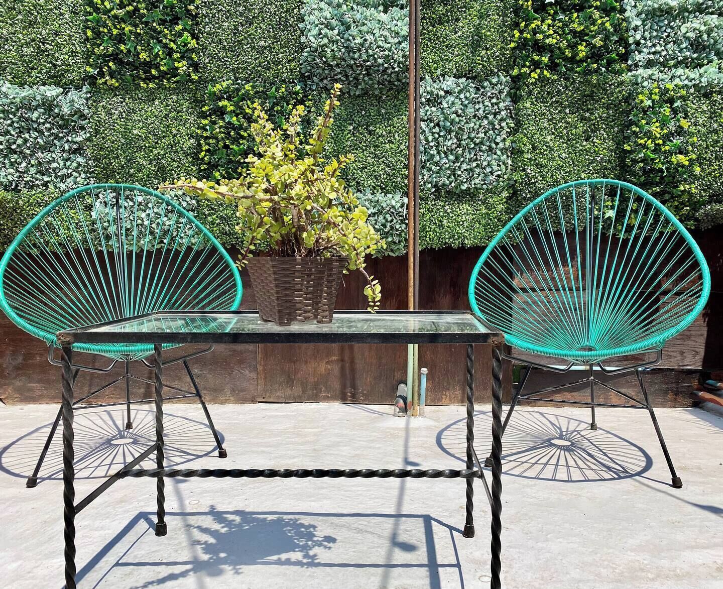 Une table avec deux chaises à armature métallique turquoise, une plante en pot et une clôture métallique en arrière-plan.