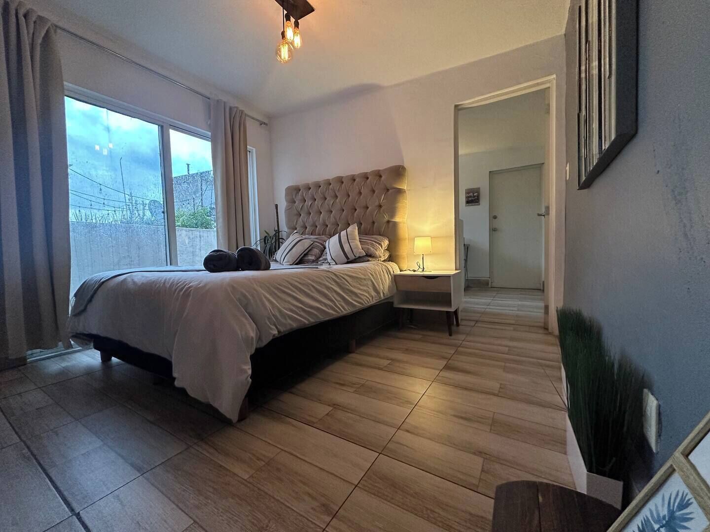 A bedroom with a large bed, a bedside table, a lamp, and a view through a window.