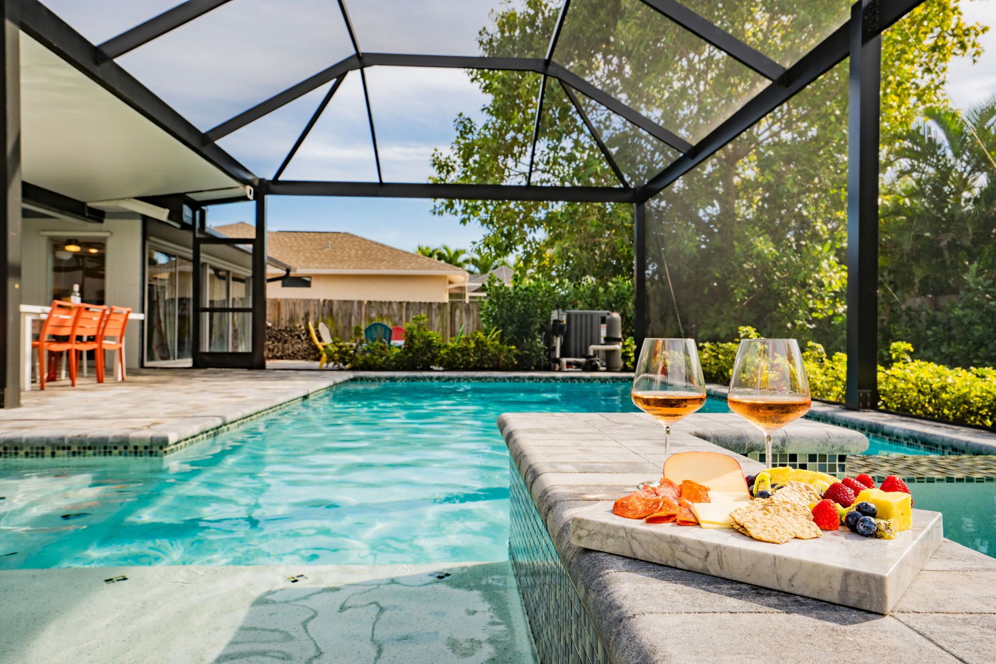 A swimming pool with a glass enclosure, a patio area with outdoor furniture, and a fruit and cheese board with glasses of wine.