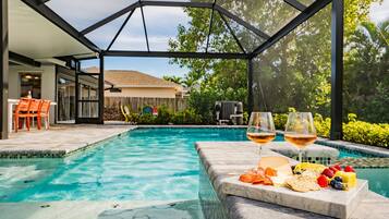 A swimming pool with a glass enclosure, a patio area with outdoor furniture, and a fruit and cheese board with glasses of wine.