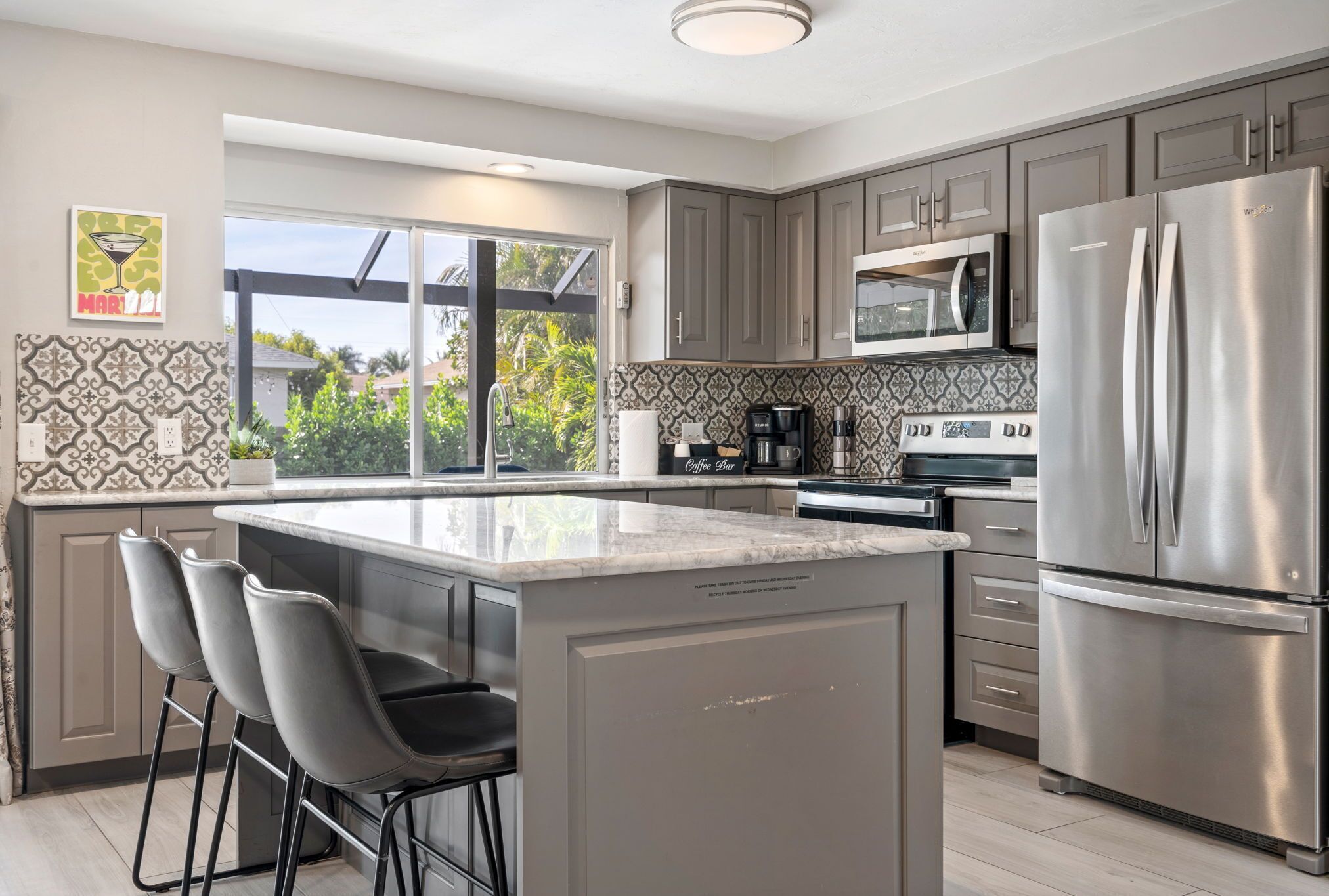 A modern kitchen with grey cabinets, stainless steel appliances, a large island with bar stools, and a view through a sliding glass door.