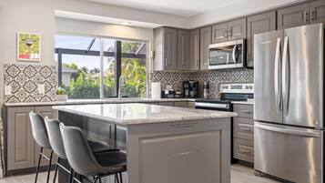 A modern kitchen with grey cabinets, stainless steel appliances, a large island with bar stools, and a view through a sliding glass door.
