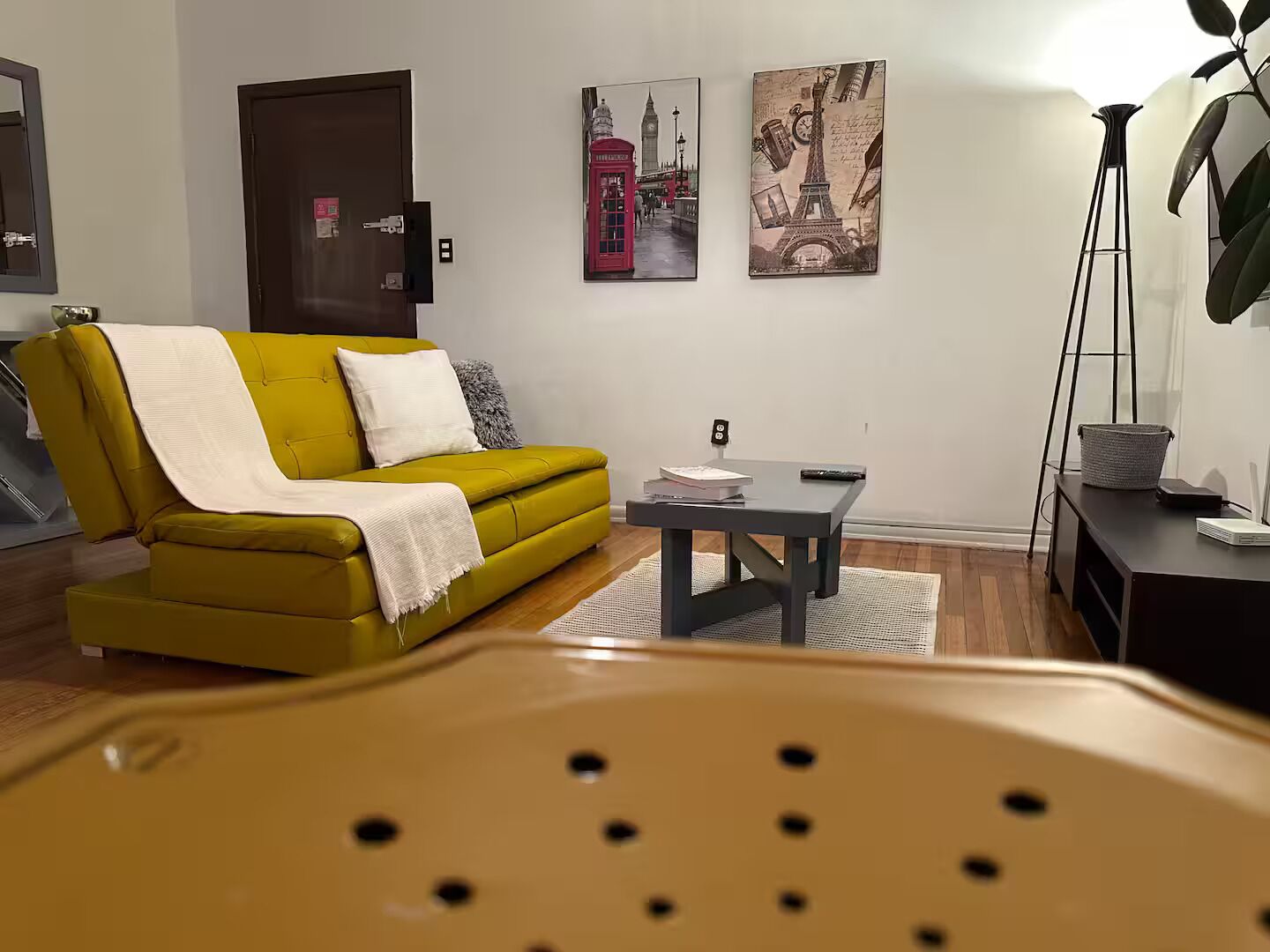 A living room with a yellow sofa, a small table, and a floor lamp. There are two paintings on the wall depicting famous landmarks.