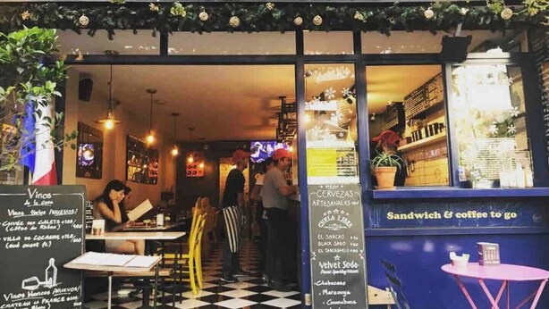 Un restaurant avec un menu écrit à la craie, des tables en plein air et une enseigne qui fait la publicité de ses sandwichs et de son café.