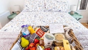 A neatly made bed with a floral bedspread, a wicker tray with fruits, snacks, and a bottle of water on the bed.