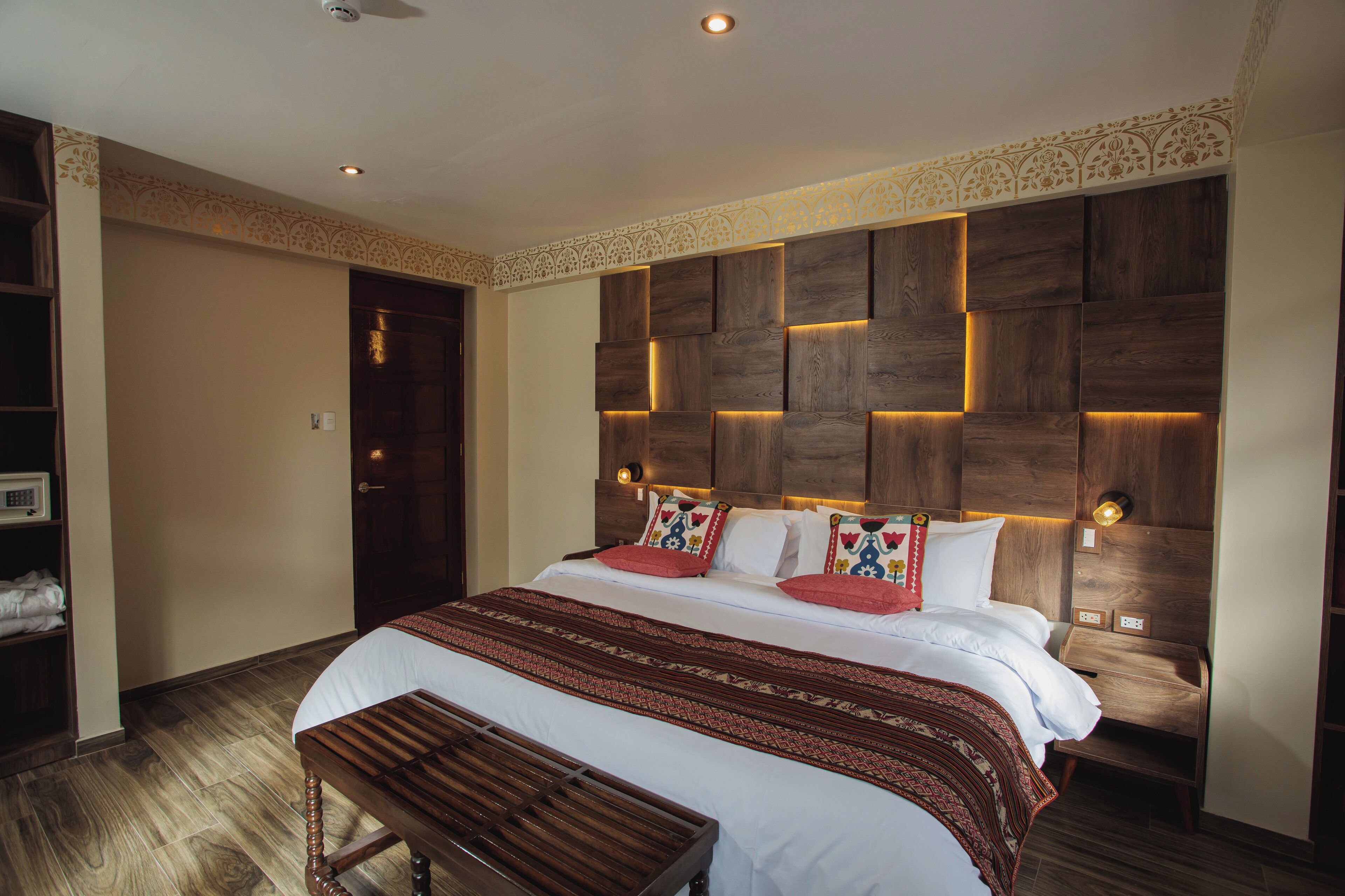 A hotel room with a large bed, a wooden bench, a wardrobe, and a decorative headboard with integrated lighting.