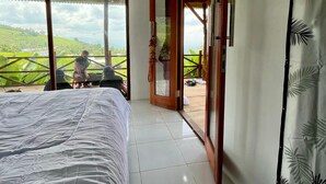 A bedroom with a large bed, a sliding glass door leading to a balcony, and a view of a green landscape.