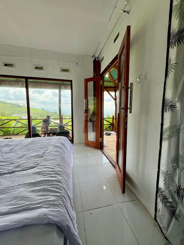 A bedroom with a large bed, a sliding glass door leading to a balcony, and a view of a green landscape.