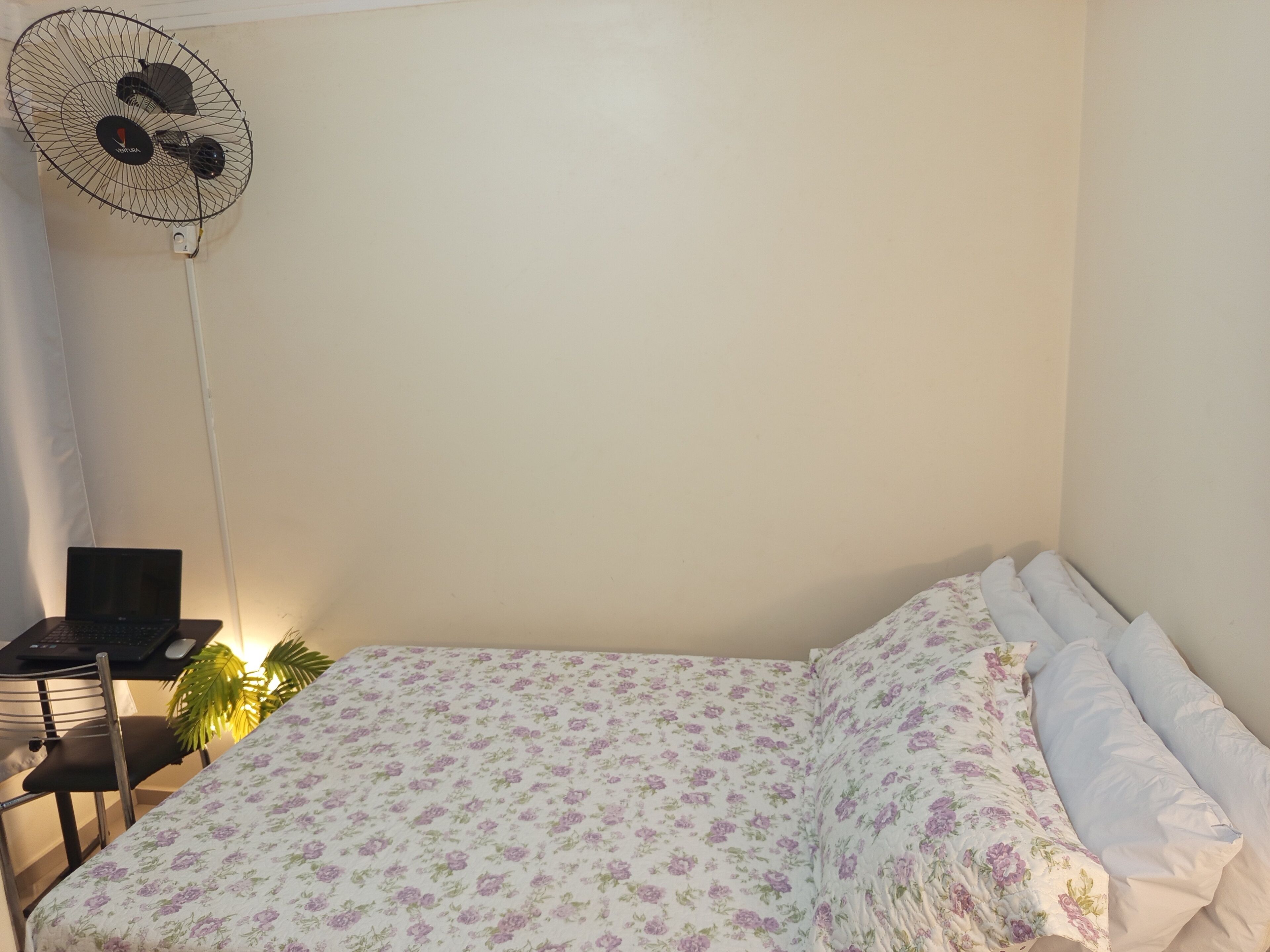 A bedroom with a bed, a desk with a laptop, and a standing fan.