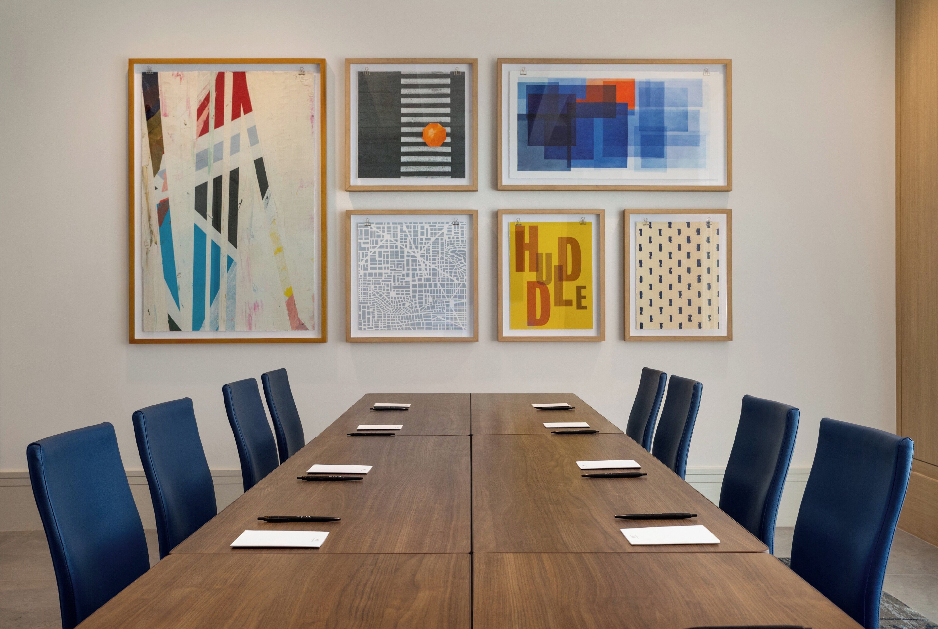 A conference room with a long table, blue chairs, and framed artworks on the wall.